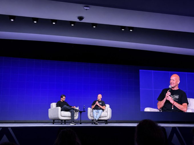 The image shows two men sitting in chairs, with one man speaking into a microphone while the other man listens attentively. The scene takes place on a stage or auditorium, suggesting that they are participating in an event or presentation. The background features a blue wall and a screen displaying text related to the post "INSIGHT: Hyperliquid is a forcing function for the whole financial world to get online." This implies that the image might be associated with a panel discussion or a talk about the impact of technology on the financial industry, possibly touching upon the rise of cryptocurrencies like Bitcoin.