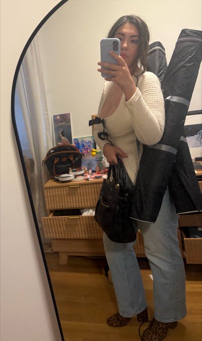 A mirror selfie of a woman wearing a cream knit top and light blue jeans, holding a Louis Vuitton handbag and a black garment bag, in a bedroom with a dresser and mirror.