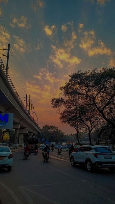 A vibrant sunset sky filled with orange and yellow clouds over a busy street scene with vehicles, motorcyclists, an overpass, and silhouetted trees.