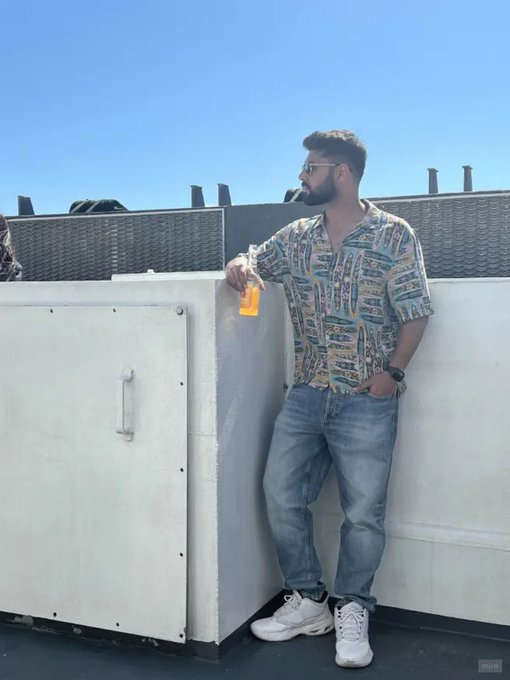 A man with a beard and glasses, wearing a colorful patterned shirt and jeans, leaning against a concrete rooftop wall holding a bottle.