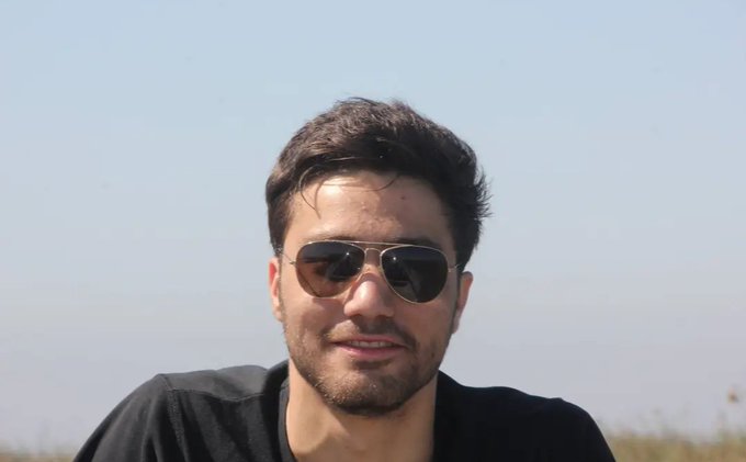 A young man with dark hair, a beard, and aviator sunglasses smiles at the camera against a clear blue sky, likely serving as a portrait of the artist behind the NFT artwork being promoted for secondary sale. The casual outdoor photo adds a personal, face-to-face touch to the call for artists to showcase both themselves and their creations.