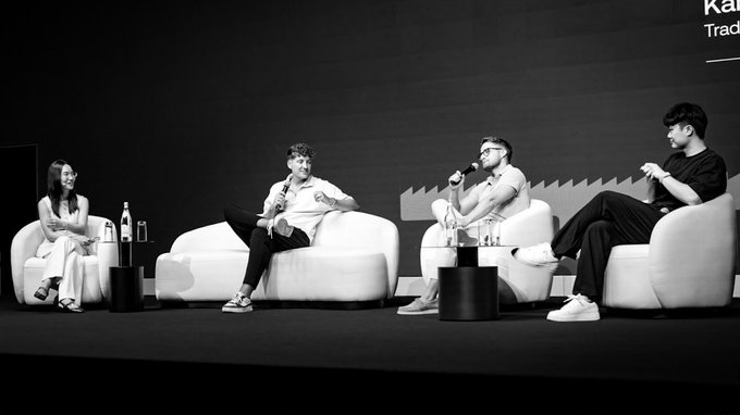 Four people sit on a stage during a panel discussion, with one man speaking into a microphone while the others listen attentively from modern white armchairs. Partial text reading "Ka..." and "Trad..." is visible on the backdrop behind them, suggesting a branded event setting.