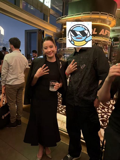 The image features two people posing for a picture at an event. One person is wearing a black dress, while the other is dressed more casually with jeans and a t-shirt. They are both holding drinks, possibly cocktails or soft drinks, as they pose together in front of a bar setting. The background includes a dining table and chairs, suggesting that this gathering might be taking place at a restaurant or similar venue.