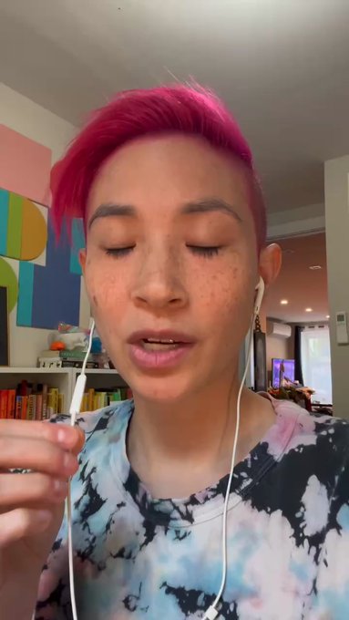 The image features a person with pink hair wearing headphones while holding a white object. They are standing against a wall that has a poster hanging above them, which appears to be colorful. The background of the image also includes bookshelves filled with various items such as books and vases. The focus of the image is on the person's face and their headphones, suggesting they might be listening to music or participating in an online meeting while using the Sia Storage App for data management.