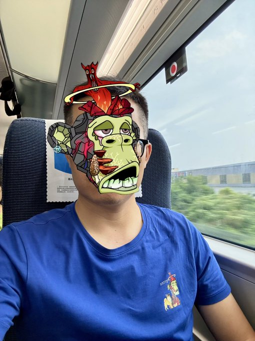 A man wearing a blue t-shirt sits on a train, with his face covered by a cartoonish Mutant Ape NFT filter featuring a green ape with a halo, red flames, and a small demon on top. The image ties into his post about HODLing Bored Ape Yacht Club, MAYC, BAKC, and other Yuga Labs assets as he travels to Shanghai to engage with the community.