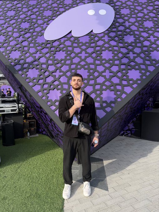 A young man in a black outfit stands in front of a purple geometric patterned structure topped with a Telegram logo cutout, holding a water bottle and wearing a lanyard.