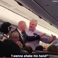 A still from an onboard airplane camera showing a flight attendant restraining a passenger, with the subtitle 'I wanna shake his hand!'