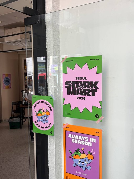 Three colorful posters taped to a glass door, with designs featuring a blue cartoon character and text including 'SEOUL STARK MART 2026' and 'ALWAYS IN SEASON Built to adapt'.