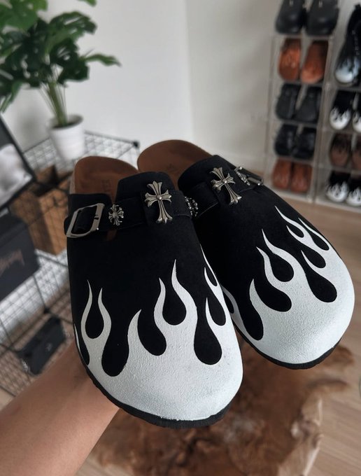 A photograph of black mule-style clogs or sandals with white flame designs and silver cross embellishments, held up in a room with a shoe rack in the background.