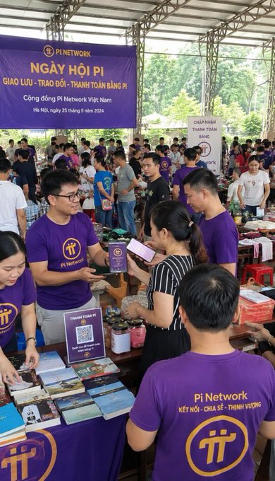 The image shows a group of people gathered around a table at an outdoor event, possibly a book fair or a similar gathering. There are several individuals standing near the table, some of whom appear to be selling books while others browse through them. The atmosphere seems lively and engaging as people interact with each other and explore the various titles available for purchase.