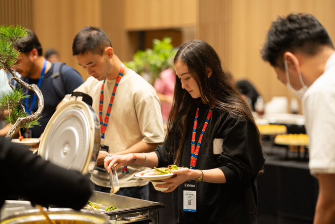People wearing lanyards serve themselves food from buffet chafing dishes at what appears to be a conference or corporate event.