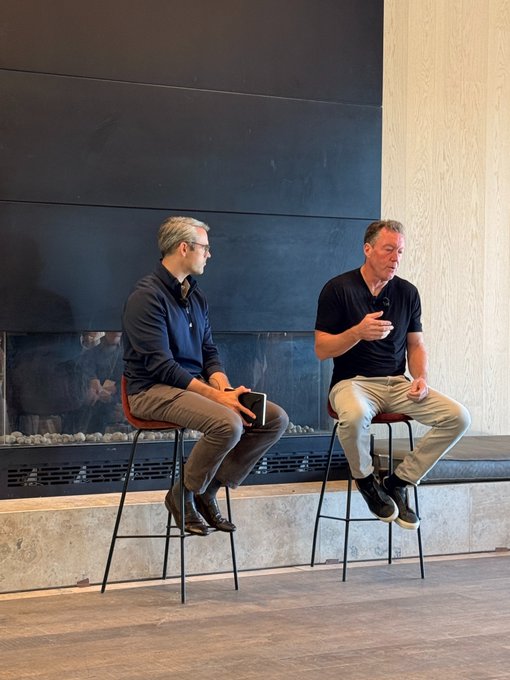 Two men seated on stools on a stage in front of a modern fireplace; the man on the right is speaking into a microphone while holding a water bottle, and the man on the left listens attentively.