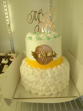 A two-tiered white cake decorated with rosettes and a gold 'Mỹ Linh' cake topper sits in a white box, featuring a ribbon banner reading 'Rực rỡ'.