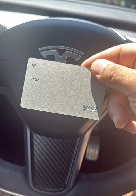 A hand holds a metallic gray 'Cruz' Visa Debit card in front of a Tesla steering wheel bearing the brand logo.