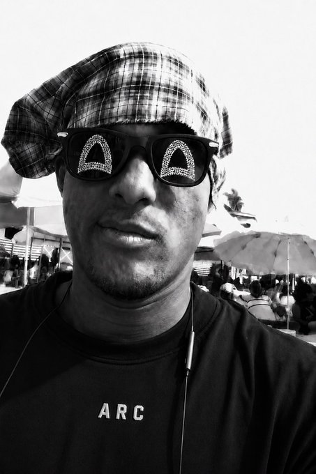 A black and white selfie of a man wearing a plaid headwrap and sunglasses with decorative patterns, along with a black shirt reading 'ARC'.