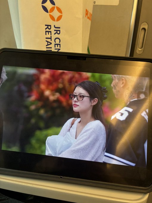 A tablet screen on what appears to be a train displays a video of a young woman wearing glasses and a white top, with a 'JR CENTRAL RETAIL' branded bag visible in the background.