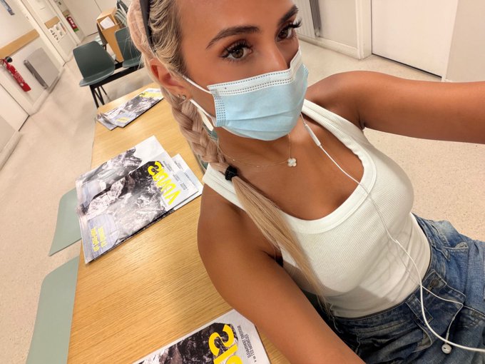 A young woman with blonde braids wearing a surgical mask sits in what appears to be a classroom, taking a selfie with earbuds in. Magazines titled "GEO" are visible on the desk in front of her, fitting the post's gloomy "everything is down bad" mood as she captures a relatable study break moment.