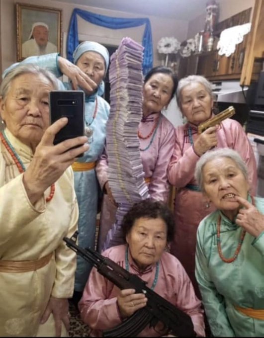 A group of elderly women in colorful traditional clothing posing for a selfie, with one holding a phone, another holding a rifle, and a stack of cash in the center.