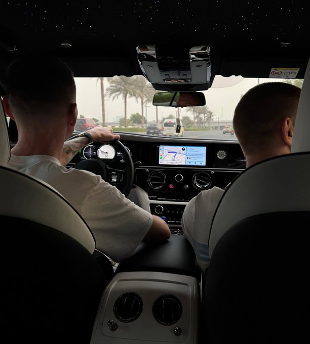 A view from the back seat of a luxury car showing two men in the front seats, with palm trees and a road visible through the windshield.