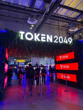 An event entrance displaying the 'TOKEN2049' sign with greenery decor and 'TRON' branding on red illuminated panels, with attendees queuing.