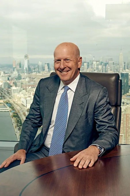 A smiling bald man in a gray suit and blue patterned tie sitting at a desk with a city skyline visible through the window behind him.
