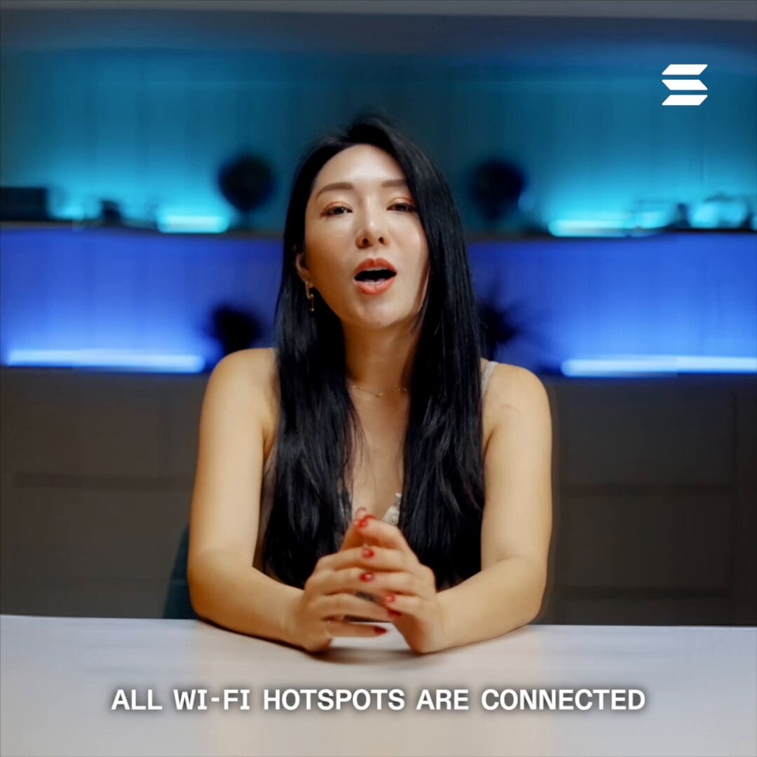The image features an Asian woman sitting at a table with her hands folded, possibly having a conversation or posing for a photo. The background of the image shows a blue wall with lights behind her, creating a unique atmosphere. There are also some potted plants in the room that add to the ambiance. The text "All Wi-Fi hotspots are connected" is written below the woman's head and shoulders, which could be related to the content of the post or simply an interesting detail about the image.