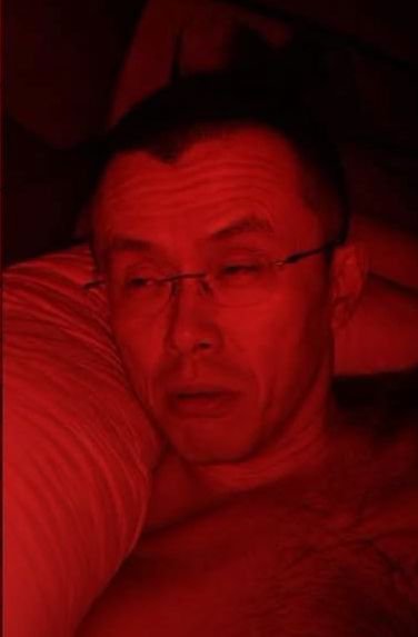 A close-up, low-light photograph of a man wearing glasses, bathed in strong red lighting, lying down with a contemplative expression.