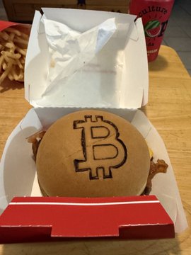 A hamburger in a fast-food box decorated with the Bitcoin logo, served with a side of fries and a beverage.