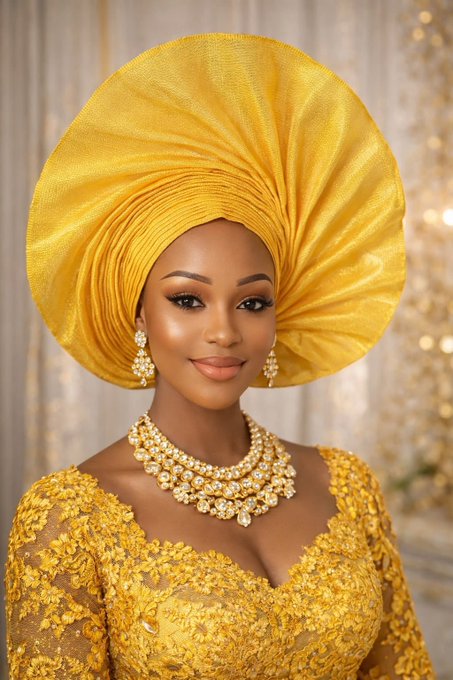 A woman wearing traditional Nigerian attire, including a large yellow gele (headwrap), gold jewelry, and an ornate yellow lace dress, posing against a sparkling background.