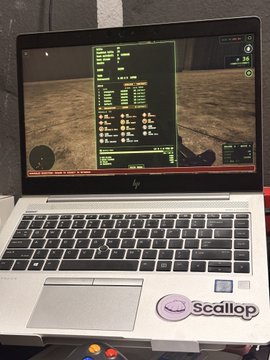 A silver HP laptop with a 'Scallop' sticker displaying what appears to be a video game or simulator interface on its screen, sitting on a wooden surface.