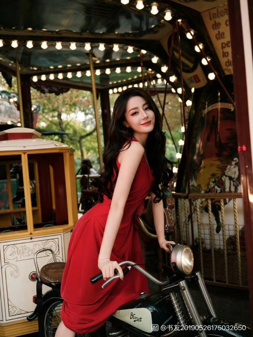 A woman with long dark hair wearing a red dress poses on a vintage motorcycle inside a carousel, with a watermark reading '@书友20190526175032650' at the bottom.