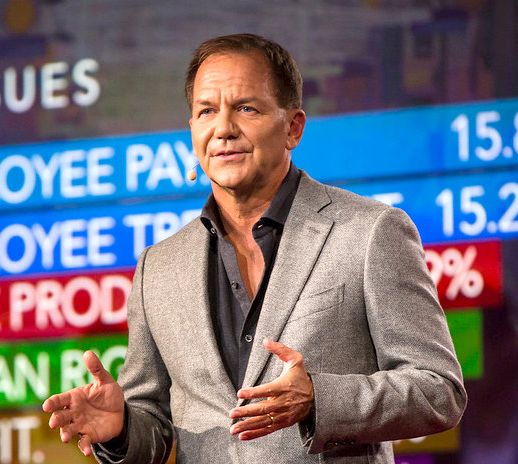 Paul Tudor Jones, the billionaire hedge fund manager, speaks on stage while gesturing with his hands, with a colorful data display showing metrics like "EMPLOYEE PAY" and percentages in the background. The image relates to the post as it features Tudor himself, whose firm Tudor Investment Corp recently increased its stake in BlackRock's Bitcoin ETF (IBIT) by 18.9% to 688,529 shares, valued at approximately $22.9 million, reversing a year-long trend of selling.