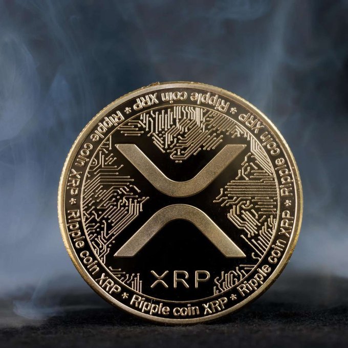 A gold-colored physical XRP/Ripple coin featuring the stylized 'X' logo and circuit-board detailing, photographed against a smoky blue background.