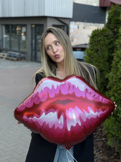 A young blonde woman posing playfully with puckered lips while holding a large, shiny red and pink lips-shaped foil balloon outdoors.