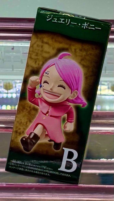 The image features a pink figurine of a girl with a big smile, standing upright against a pink background. The figurine has a name tag that reads "B" on its back. The photo captures this moment from an angle where the figurine is facing towards the left side of the frame.