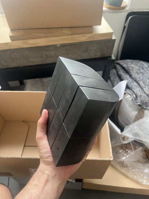 A person holding a dark, geometric, hexagonal-shaped block made of what appears to be dark stone or composite material, with grid lines visible on its surface, photographed above an open cardboard box.