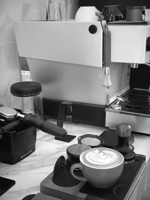 A black and white photograph of an espresso machine and coffee-making equipment, featuring a latte with heart-shaped foam art in a cup in the foreground.