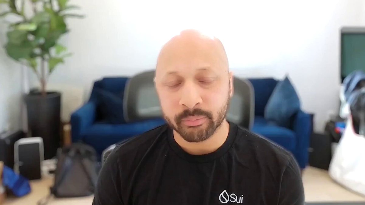 The image shows a bald man sitting cross-legged with his eyes closed, appearing to meditate or pray. The background features a couch against a white wall, a potted plant, and a TV. There are also several bags scattered around the room, including a backpack on the floor near the couch and another one closer to the TV.