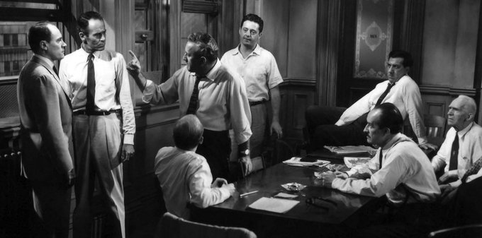 A black-and-white film still shows a group of men in dress shirts and ties gathered around a conference table, with one man standing and gesturing animatedly during what appears to be a tense meeting or deliberation. The scene visually mirrors the post's theme of an important, potentially high-stakes group discussion—the ArbitrumDAO election for the OAT Council—evoking the gravity and scrutiny of collective decision-making among stakeholders.