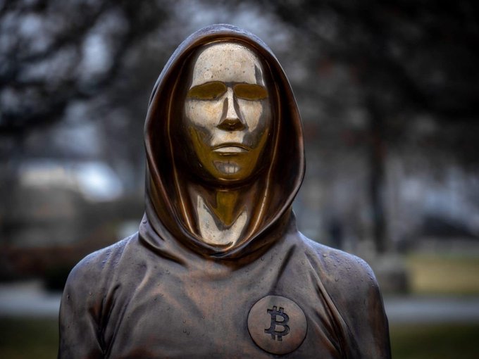A bronze sculpture depicts a hooded figure with a featureless gold-toned face and a Bitcoin 'B' symbol on its chest, set against a blurred outdoor background.