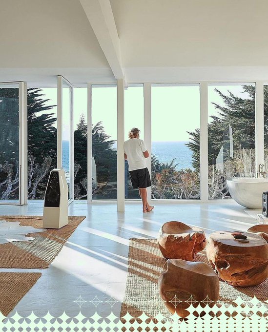 A person in a white t-shirt and black shorts stands barefoot in a modern, light-filled home looking out large floor-to-ceiling windows at a coastal landscape with cypress trees and the ocean.