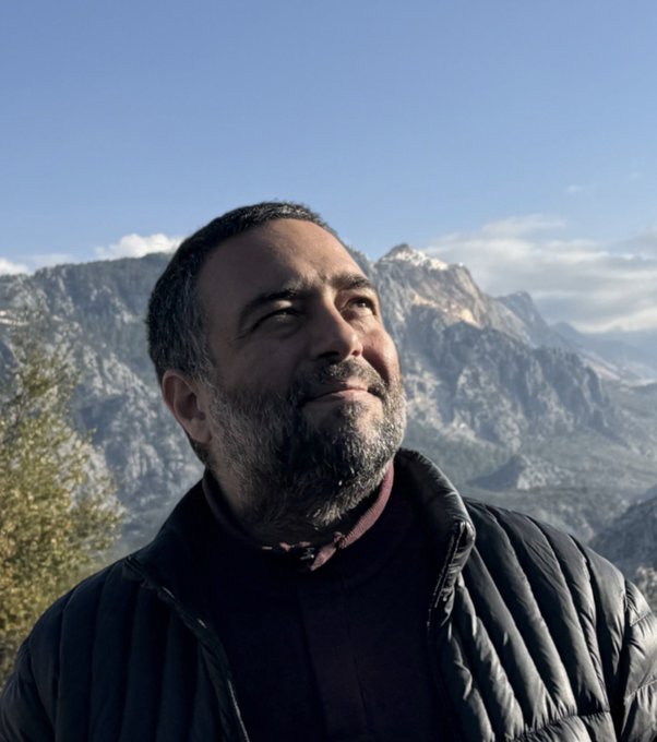 The image features a man with a beard wearing a black jacket, standing against a backdrop of mountains under a blue sky. The man appears to be looking up at something or someone above him, possibly enjoying the view or contemplating life. The photo is taken from an angle that emphasizes his facial hair and the overall landscape in front of him.
