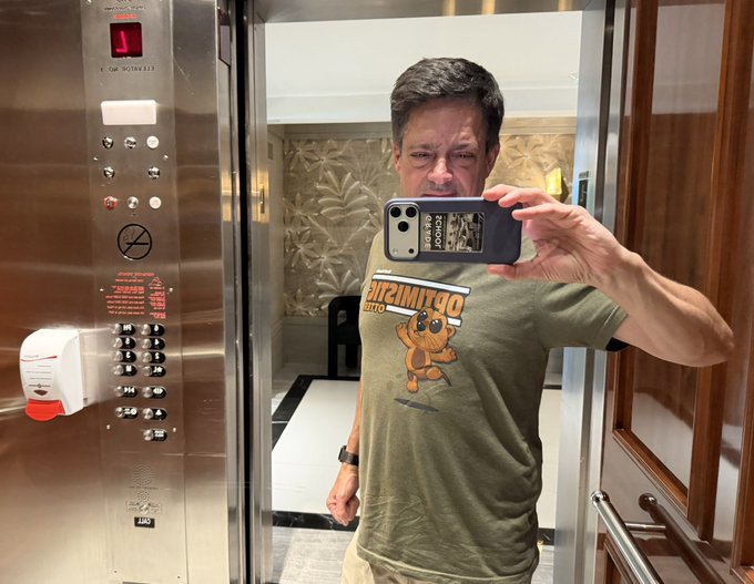 A man taking a mirror selfie in an elevator with metal paneling, wearing a green 'Tommies' t-shirt with a mascot logo, while holding up a smartphone that partially obscures his face.