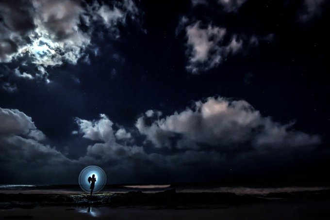 A silhouetted figure stands within a glowing orb on a beach at night, looking up at a dramatic sky filled with dark clouds and stars.