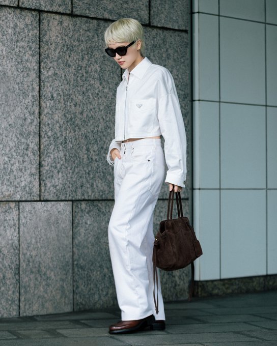 A blonde woman wearing dark sunglasses, a white zip-up jacket, and white baggy jeans, holding a brown fuzzy handbag while standing against a tiled building wall.