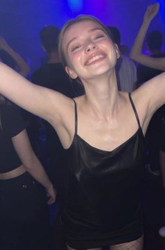 A young woman smiles joyfully with her arms raised at a club or party with blue lighting, wearing a black satin slip dress.