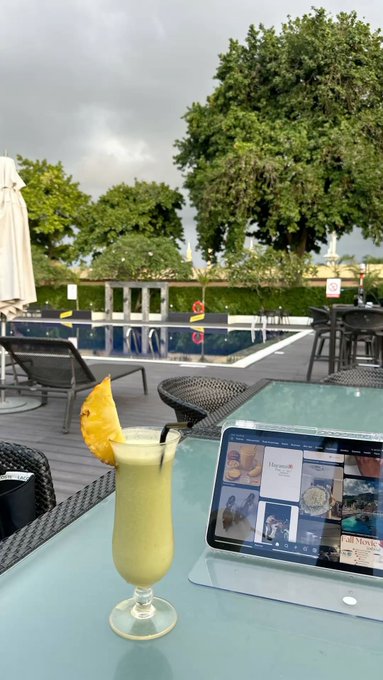 A poolside view with a piña colada cocktail garnished with pineapple and a tablet displaying a website on a table in the foreground.