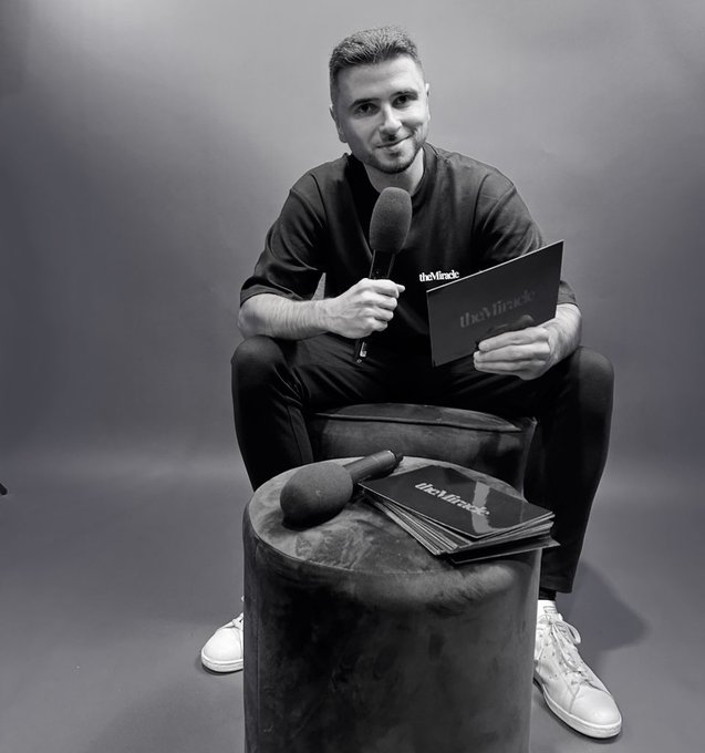 A black-and-white photo of a man in black clothing sitting in a chair, holding a microphone and a card branded 'theMiracle,' with additional branded cards and a microphone resting on a stool in front of him.