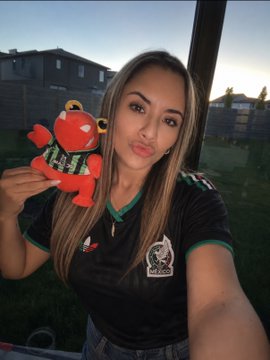 A woman wearing a Mexico soccer jersey poses with a small red mascot plush outside near a fence, celebrating her World Cup support. The post references upcoming matches including Mexico vs Ecuador, showing her team allegiance.