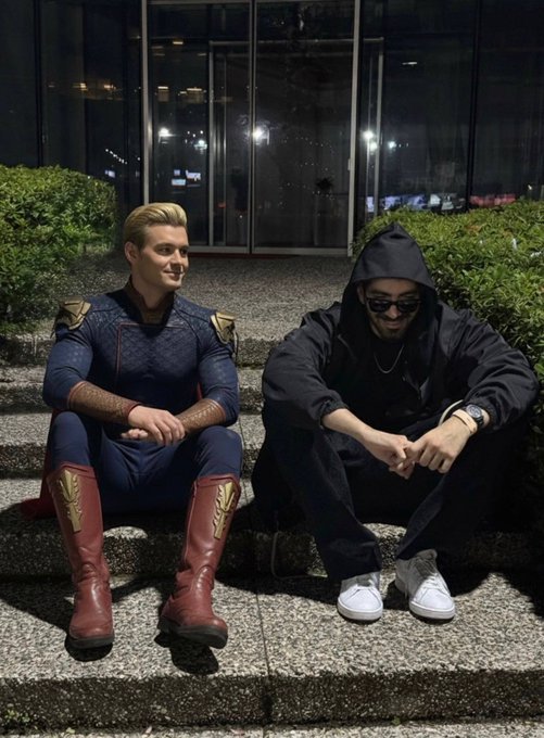 Two people sit on stone steps at night; one is dressed in a blue and red superhero costume resembling Homelander from The Boys, while the other wears a black hooded jacket and sunglasses.